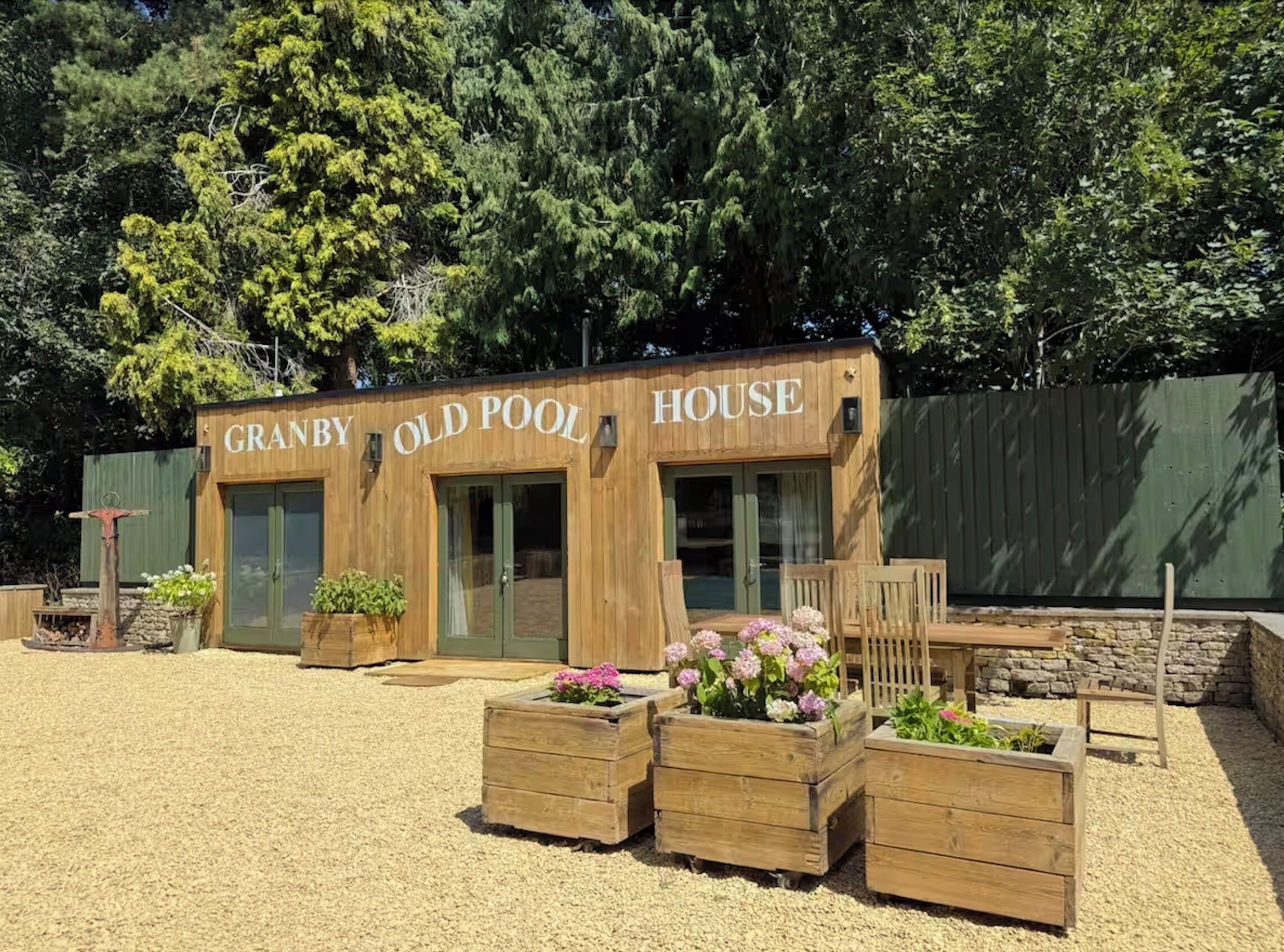 A wooden building labeled "GRANBY OLD POOL HOUSE" with a gravel ground and wooden planters with flowers.