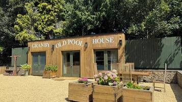 A wooden building labeled "GRANBY OLD POOL HOUSE" with a gravel ground and wooden planters with flowers.