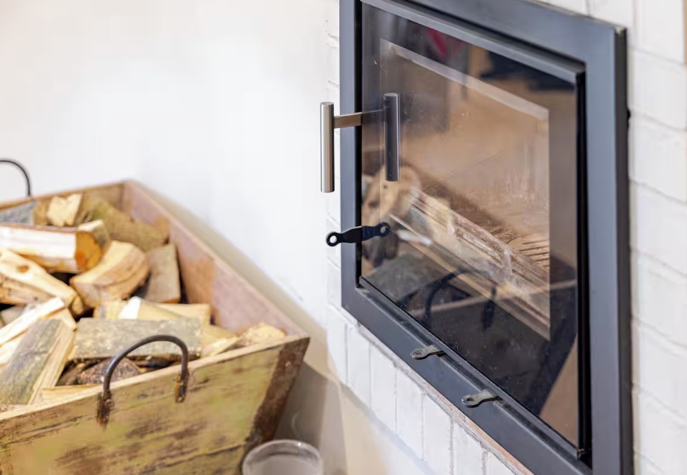 Une cheminée dotée d’une porte en verre, d’une poignée en métal et d’un coffre en bois rempli de bûches.