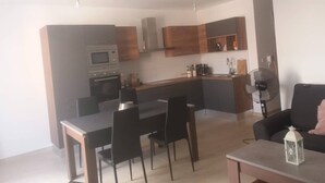 Et moderne køkken med indbyggede hårde hvidevarer, et spiseområde med bord og stole, samt en opholdsstue med sofa og ventilator.