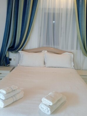 A neatly made bed with pillows and folded towels, a bedside table, and a window with sheer curtains. - Studio Flat - Sleeps 4 & Private Garden (Sarandë)
