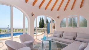 Ein modernes Wohnzimmer mit einer Couch, Sesseln und einem Glastisch. Große Bogenfenster bieten Blick auf Pool und Grünflächen.