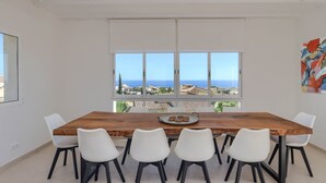 Ein Essbereich mit Holztisch und weißen Stühlen, großen Fenstern mit Meerblick und einem Deckenventilator.