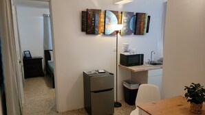 Une chambre d’hôtel compacte comprenant un coin cuisine, une poubelle, un bureau avec une chaise, une petite table, une plante en pot et une œuvre d’art accrochée au mur.