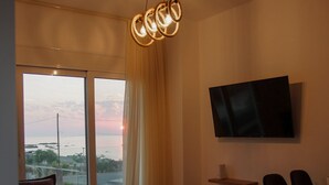 Een moderne woonkamer met een groot raam, een flatscreen televisie aan de muur, een bank, een salontafel en een eethoek met stoelen.