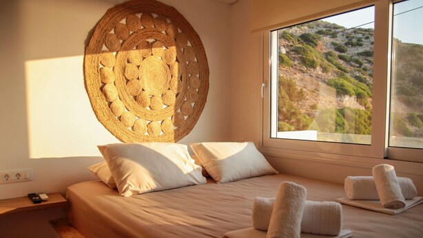 Een slaapkamer met een groot bed, twee kussens, een wanddecoratie van geweven materiaal en uitzicht op een heuvel door een raam.