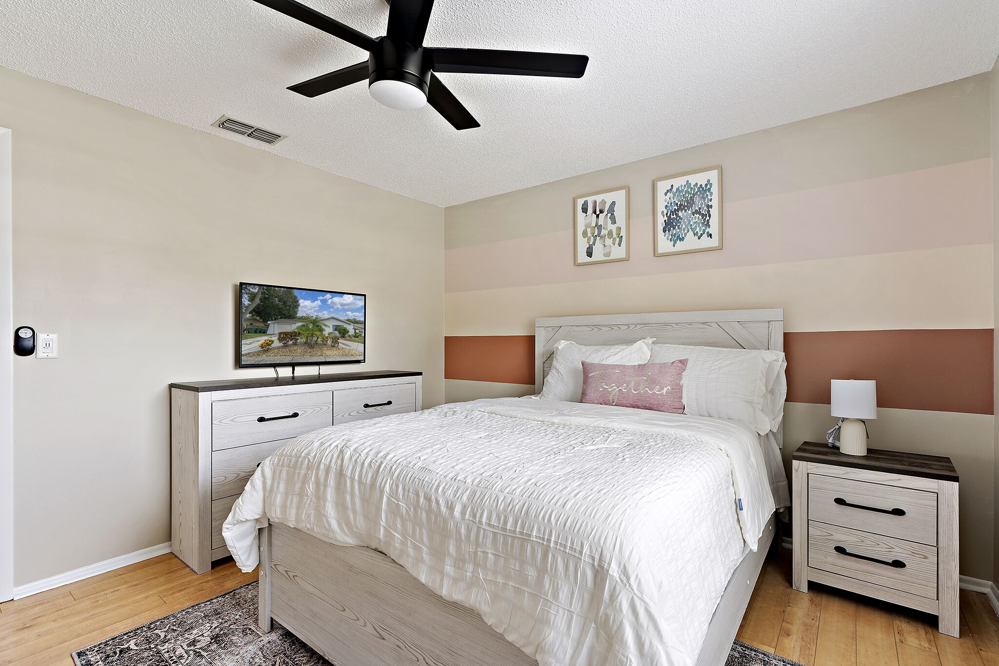 A bedroom with a bed, nightstand, dresser, ceiling fan, artwork, and a television.