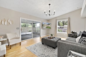 A spacious living room with a grey sofa, white armchair, and ottoman, a chandelier, and a large window with a view of a house.