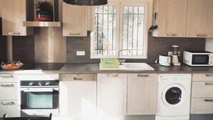 A modern kitchen with a dishwasher, oven, microwave, and washing machine. There are windows with bars, a hanging light fixture, and wooden cabinets. - Bas de Villa - Nice hinterland (Contes)