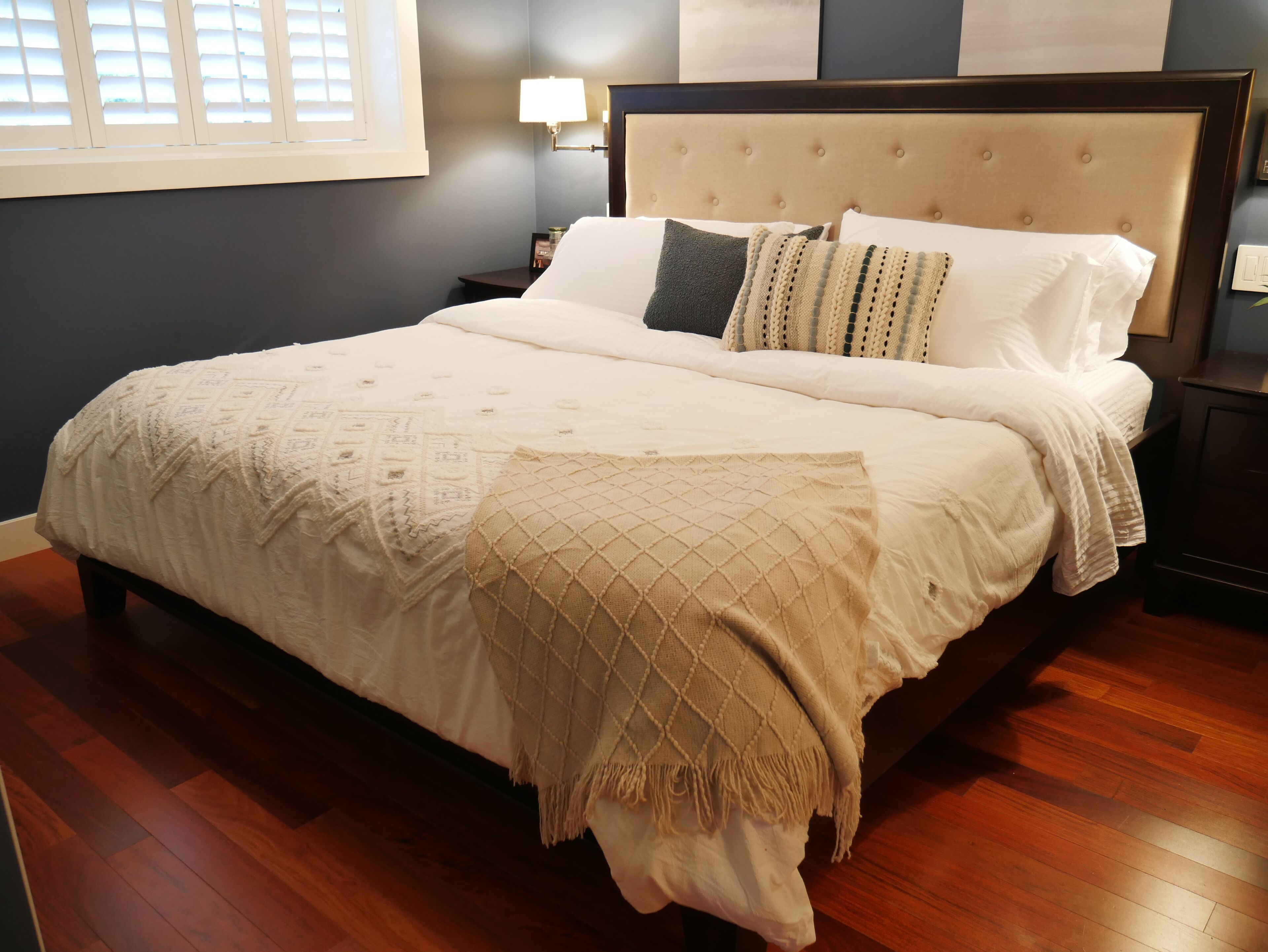 Un lit bien fait, recouvert d’une couverture beige et agrémenté d’un plaid décoratif, une table de chevet avec une lampe et une fenêtre avec des stores.
