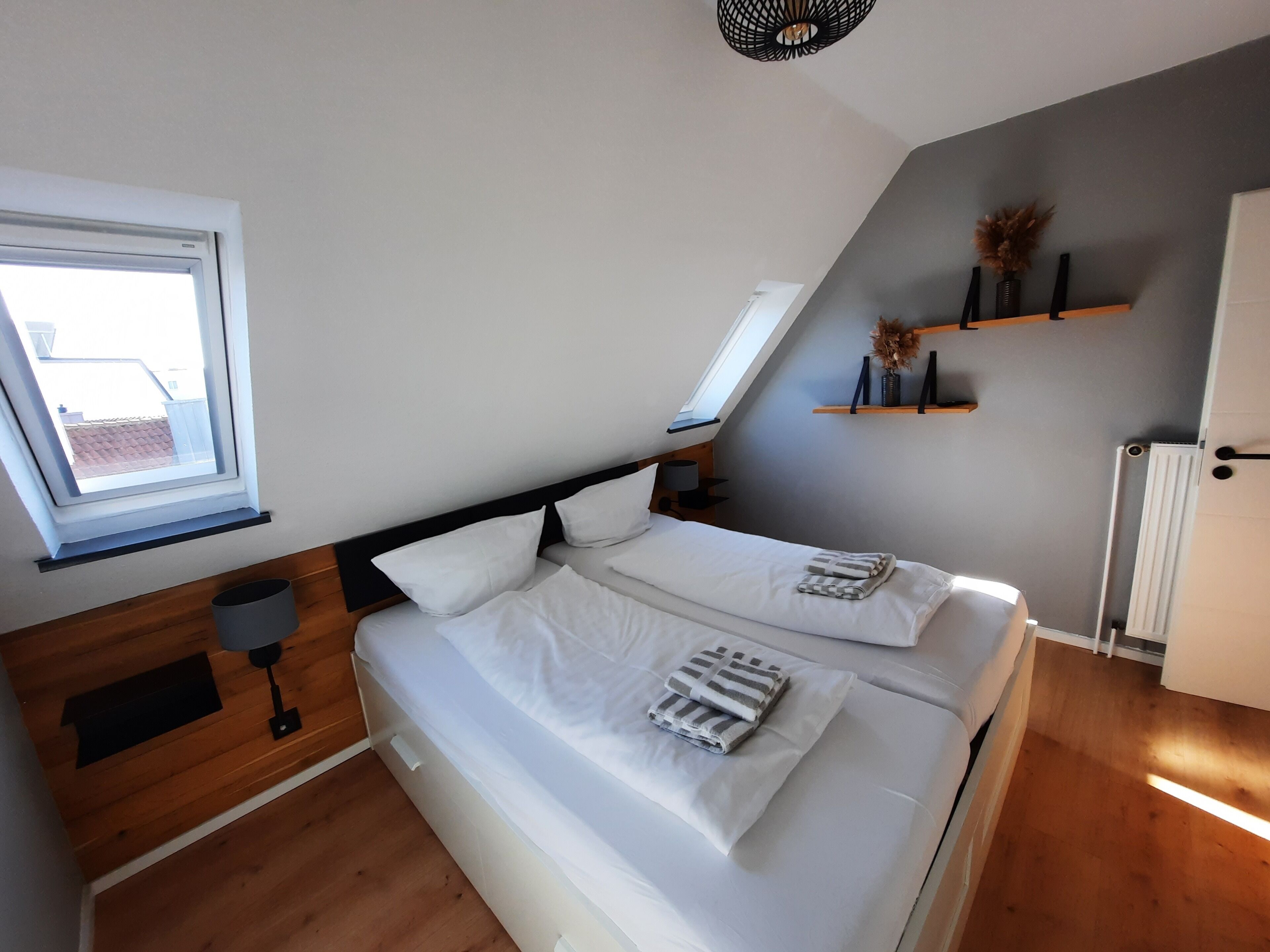 A bedroom with a sloped ceiling, a skylight, a bed with white bedding, a bedside table with a lamp, and a shelf with decorative items.