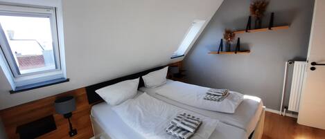 A bedroom with a sloped ceiling, a skylight, a bed with white bedding, a bedside table with a lamp, and a shelf with decorative items.