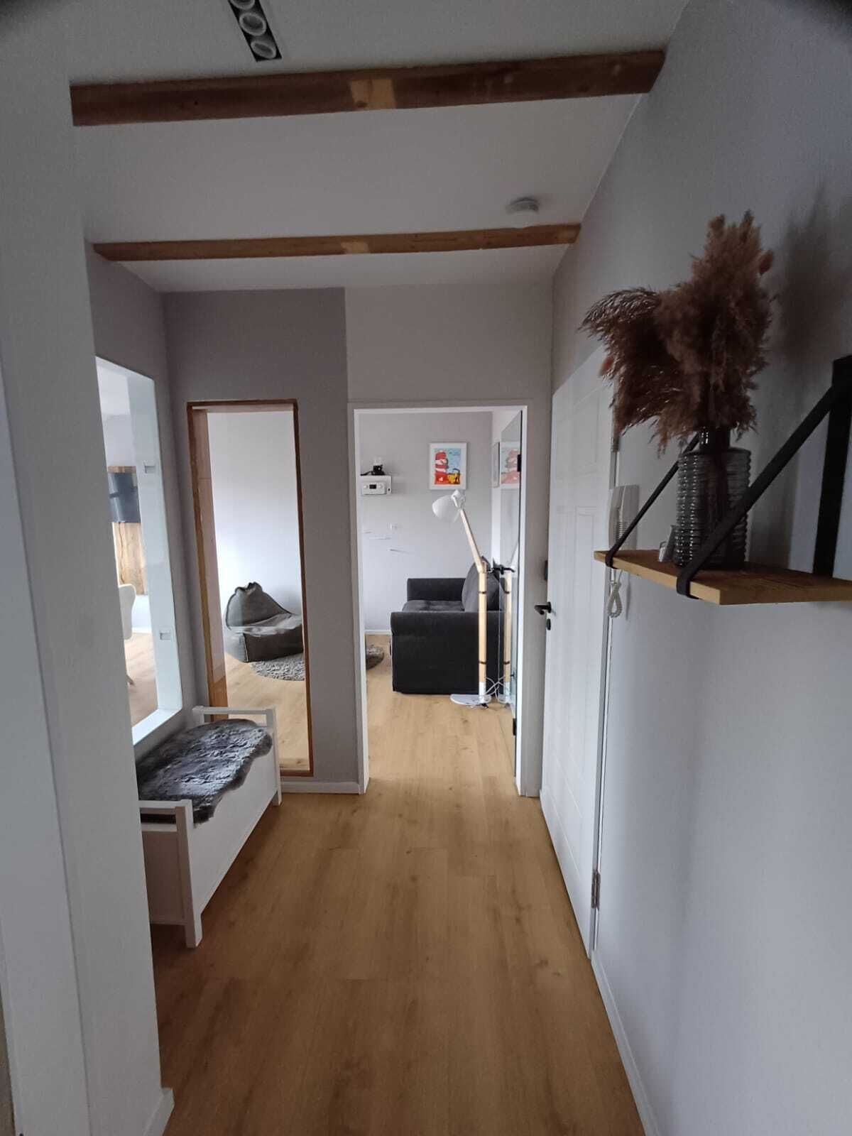 Un estrecho pasillo con piso de madera, un banco y una puerta que da a una sala de estar con sofá y lámpara.