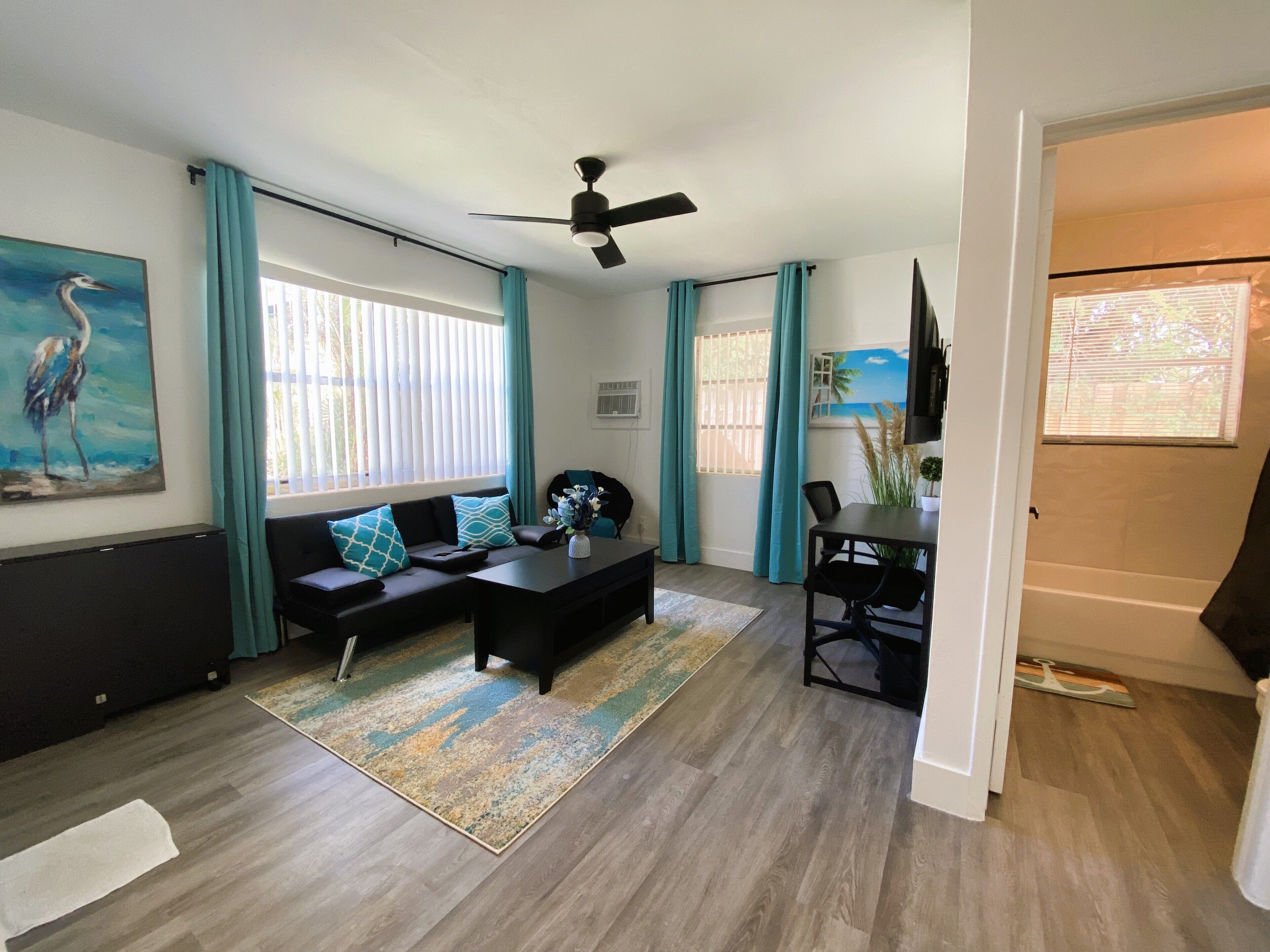 A modern living room with a sofa, coffee table, and dining table. There are windows with curtains, a ceiling fan, and a painting on the wall.