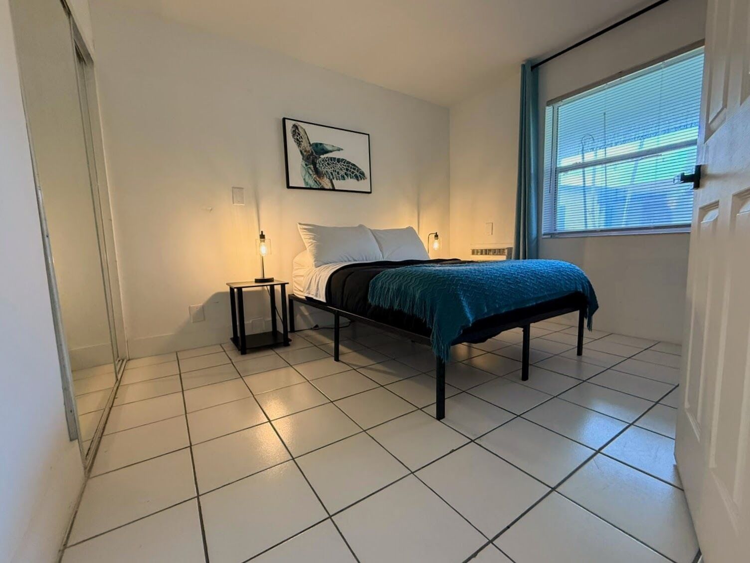 A bedroom with a bed, a nightstand, a wall-mounted light, a window with blinds, and a framed picture on the wall.