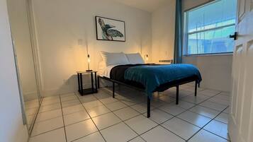 A bedroom with a bed, a nightstand, a wall-mounted light, a window with blinds, and a framed picture on the wall.