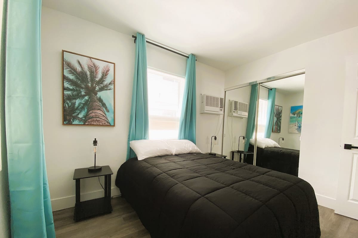 A bedroom with a large bed, a bedside table, a mirror, and a window with curtains.
