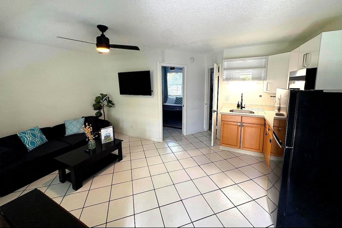 A living room with a black sofa, a coffee table, and a ceiling fan. There is a kitchen area with wooden cabinets and a refrigerator.