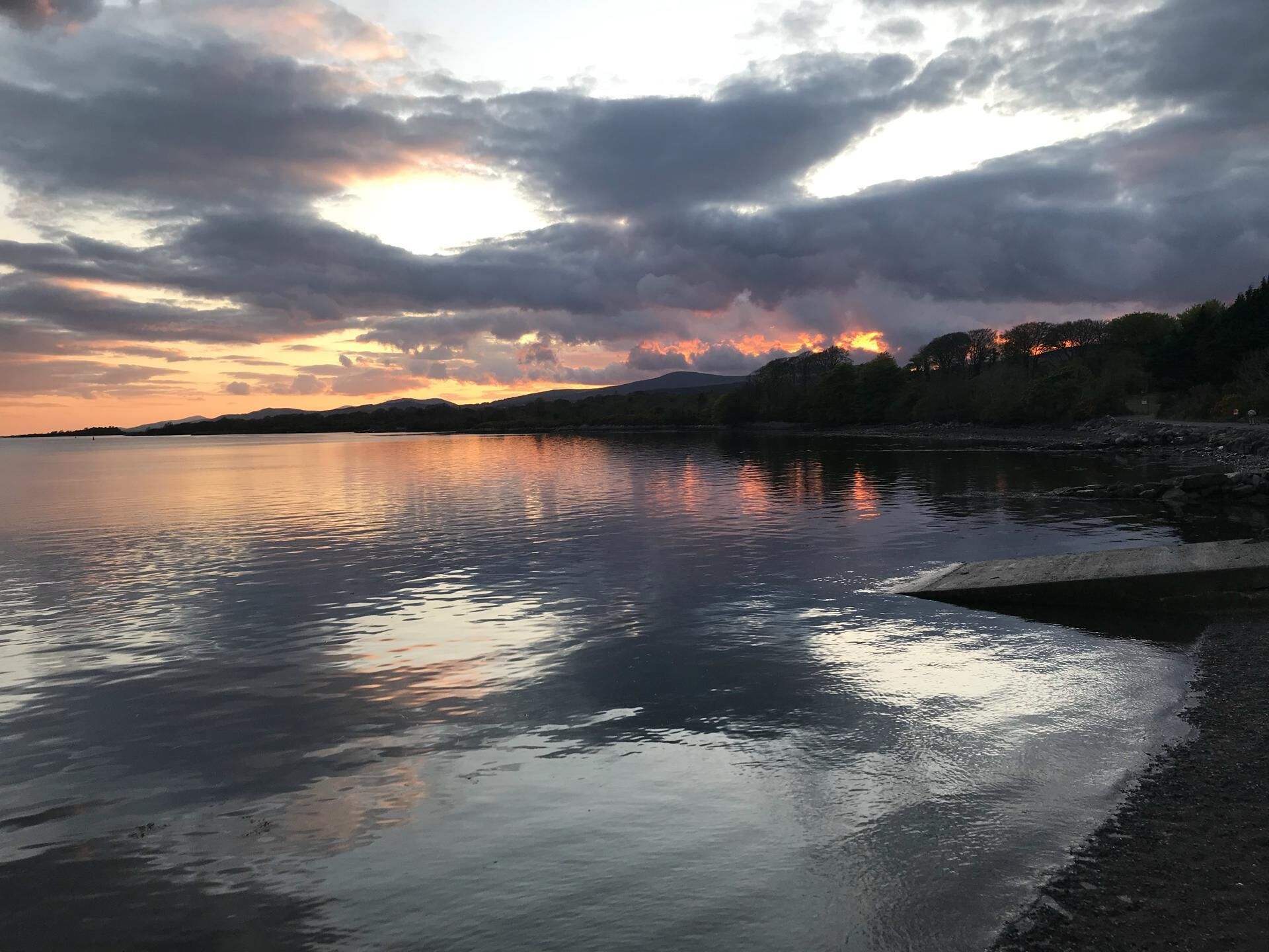 A calm lake at sunset with a rocky shoreline and distant mountains.