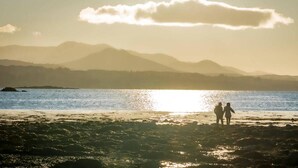 A beach with two people walking, mountains in the background, and the sun setting over the water. - Newly Renovated, Stylish Self-catering Apartment (Dawros)