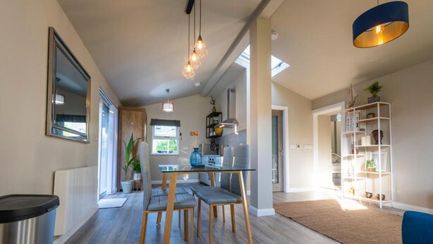 A modern kitchen with wooden cabinets, a dining area with a glass table and chairs, and a living area with a bookshelf and a blue chair. - Newly Renovated, Stylish Self-catering Apartment (Dawros)