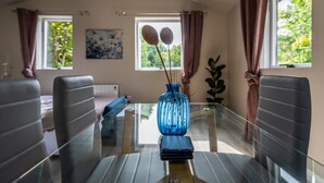 A dining area with a glass table, black chairs, a blue vase with branches, and two windows with curtains. - Newly Renovated, Stylish Self-catering Apartment (Dawros)