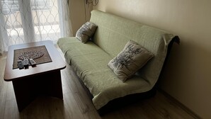 Un sofá verde con dos almohadones decorativos, una mesita de madera con un mantel tejido y controles remotos, y un televisor de pantalla plana montado en la pared.