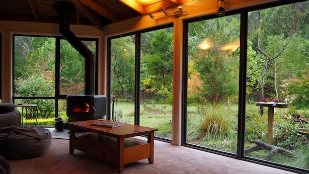 A cozy room with a fireplace, large windows, and a view of a lush garden. - Blackwood Bush Retreat (Strathewen)