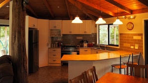 A modern kitchen with wooden beams, pendant lights, a large island, and a view of the outdoors. - Blackwood Bush Retreat (Strathewen)