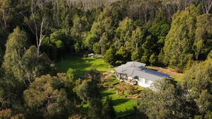 A modern house surrounded by dense forest, with a well-maintained lawn and solar panels on the roof. - Blackwood Bush Retreat (Strathewen)