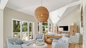 Un vaste salon doté d’un haut plafond, de grandes fenêtres et offrant une vue sur l’extérieur. On y trouve un coin repas avec une table et des chaises, ainsi qu’un téléviseur fixé au mur.