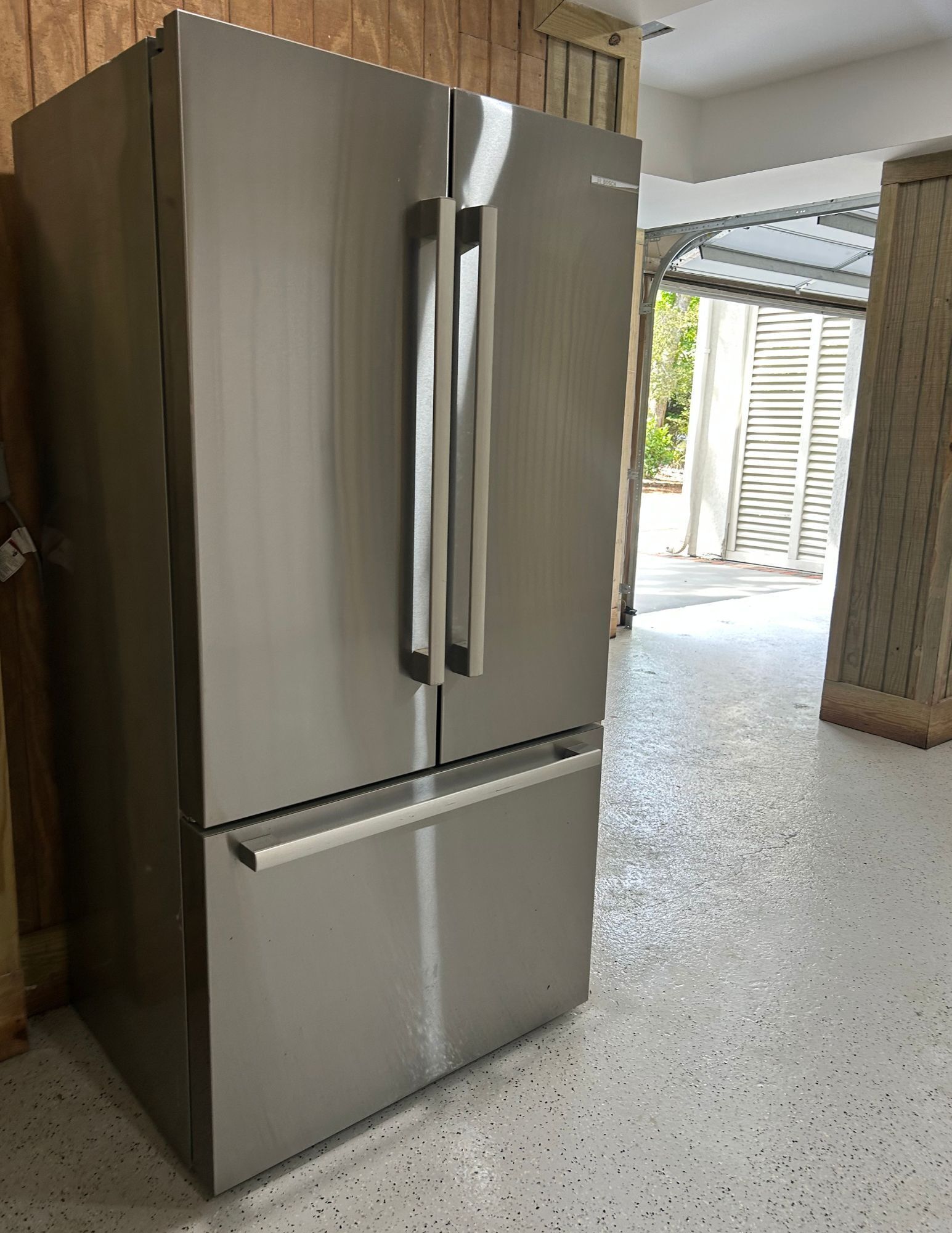 Een roestvrijstalen koelkast in een keuken met houten kastjes en een betonnen vloer.