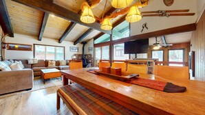 A spacious living room with a wooden dining table, benches, and a fireplace. There are large windows allowing natural light and views of the outdoors.