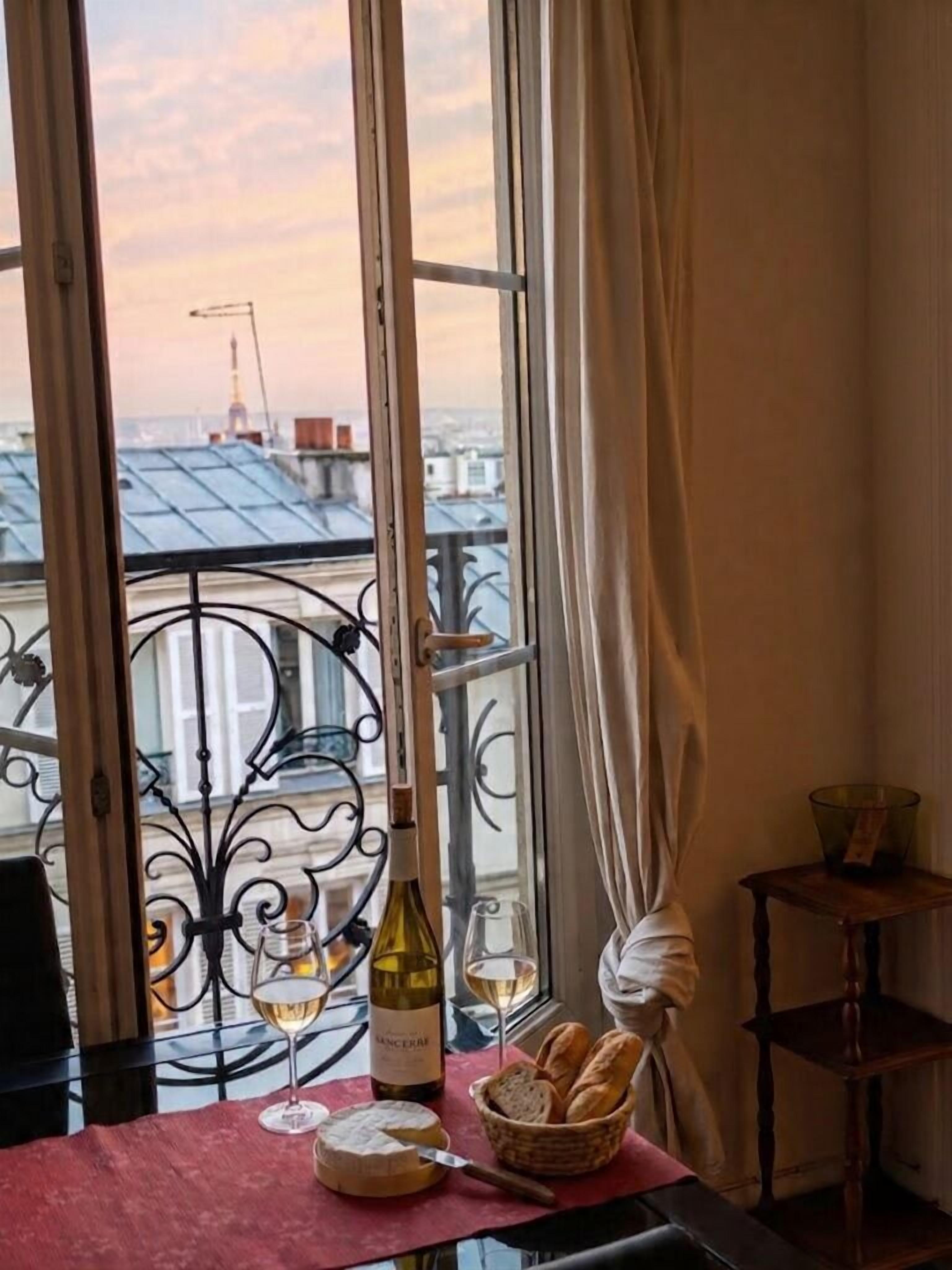 A table with a bottle of wine, glasses, a cheese wheel, and a basket of bread, set against a window with a view of buildings and a crane.