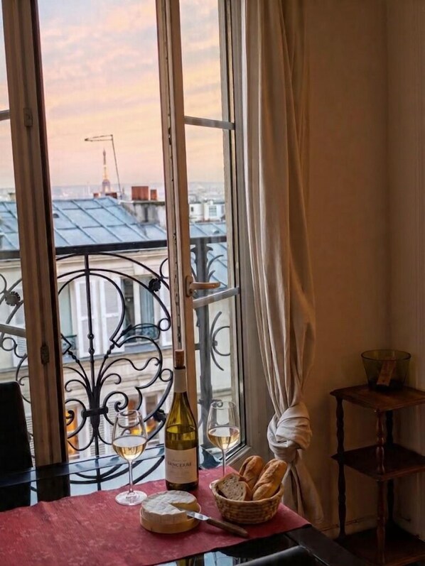 A table with a bottle of wine, glasses, a cheese wheel, and a basket of bread, set against a window with a view of buildings and a crane.