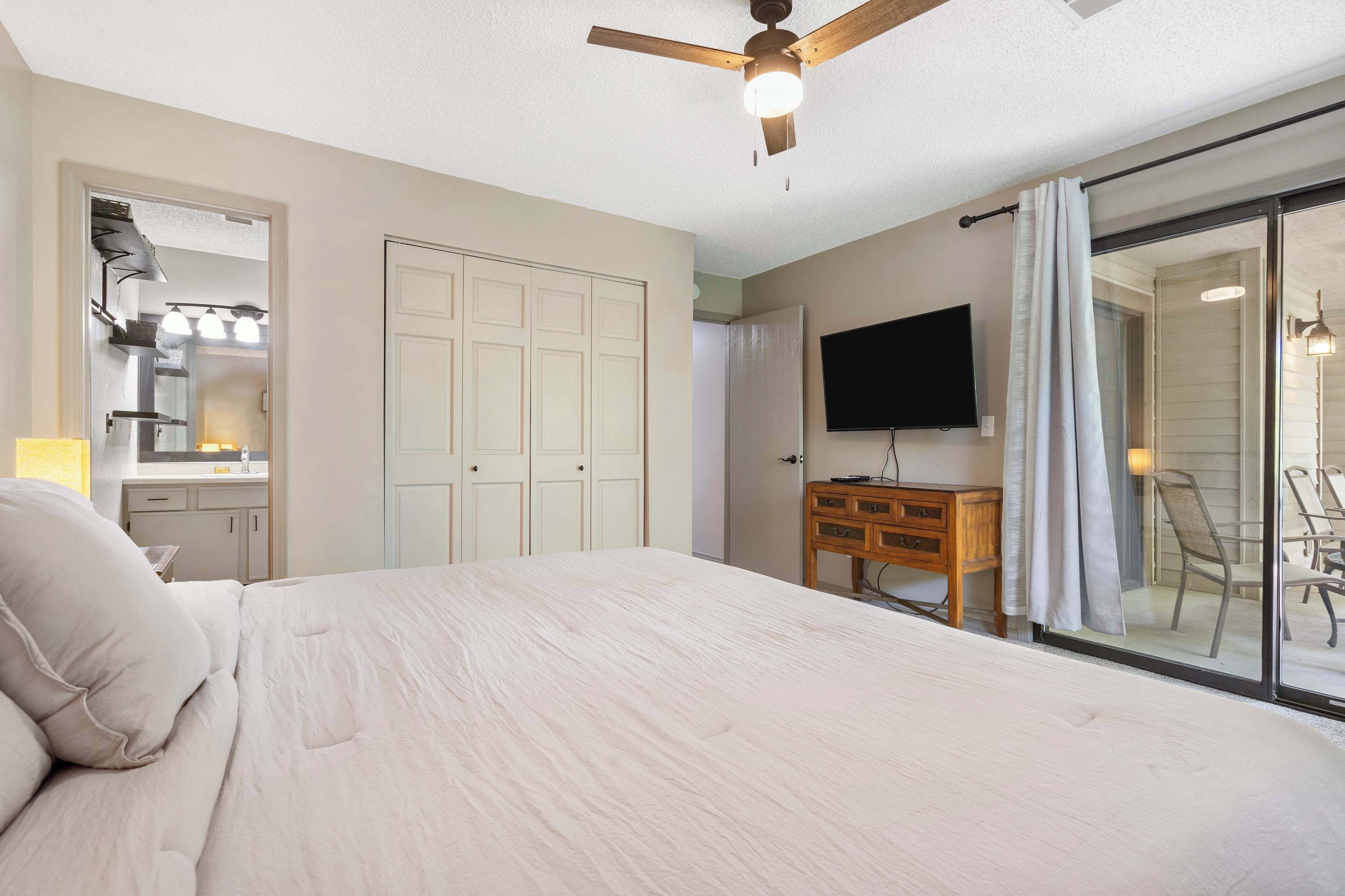 A bedroom with a bed, a ceiling fan, a TV, a wooden dresser, and a sliding glass door leading to a balcony.