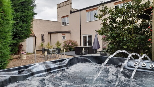 Un jacuzzi dans un jardin, avec une table et des chaises sur une terrasse, entouré de verdure et avec une maison en arrière-plan.