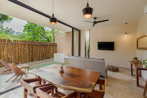 A spacious patio with a wooden table and chairs, a ceiling fan, and a TV mounted on the wall.