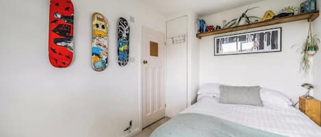 Une chambre à coucher avec un lit, une étagère murale contenant des livres et des objets décoratifs, et trois planches de skate accrochées au mur.