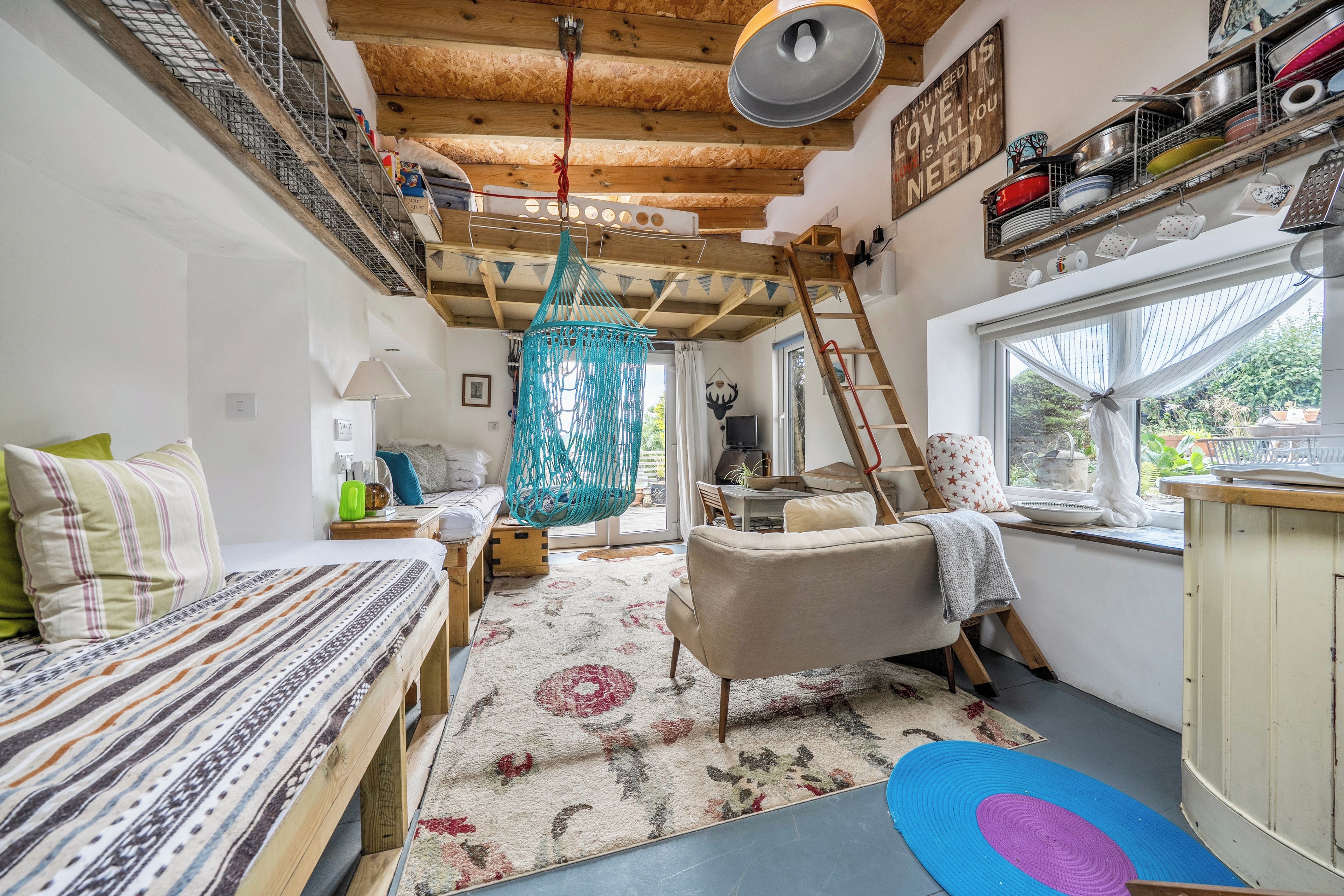 Une pièce chaleureuse comprenant un lit, un canapé, un fauteuil suspendu en macramé, une échelle en bois et une fenêtre donnant sur un paysage verdoyant.