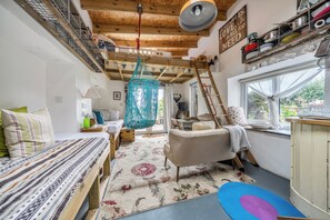 A cozy room with a bed, a sofa, a hanging macrame chair, a wooden ladder, and a window with a view of greenery.