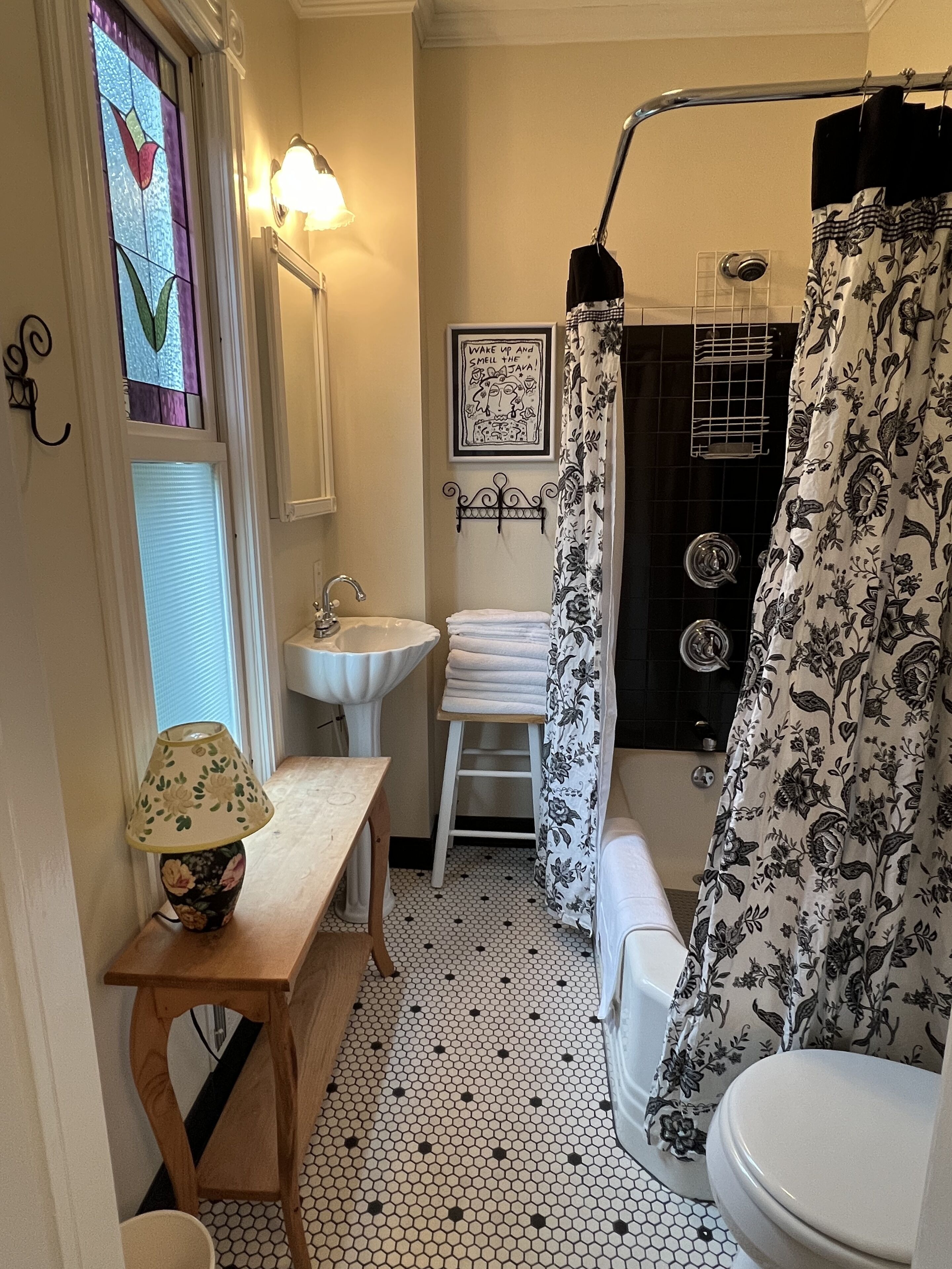 A bathroom with a sink, toilet, and shower. There is a window with a stained glass design, a wall-mounted light, and a framed picture on the wall.