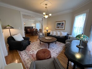 A living room with a dining area, featuring a chandelier, patterned rug, and a painting on the wall.