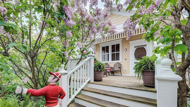 A wooden staircase leading to a porch with a white railing, surrounded by lush greenery and flowering trees. - Charming Victorian, Private Hot Tub (New Buffalo)