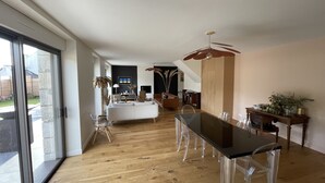 Ein geräumiger Wohnbereich mit einem großen Glasfenster, das auf einen Balkon führt, einem Esstisch mit transparenten Stühlen und einem modernen Deckenventilator.