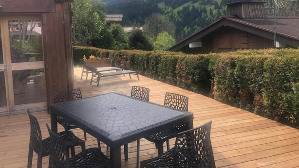An outdoor wooden deck with a table and chairs, surrounded by hedges and a mountainous backdrop. - Chalet, Ground floor, parking, tv, ski locker, 44m², Le Grand Bornand (Le Grand-Bornand)