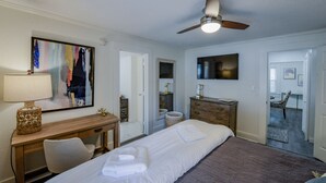 A hotel room with a bed, a desk, a chair, a ceiling fan, a TV, and a painting on the wall.