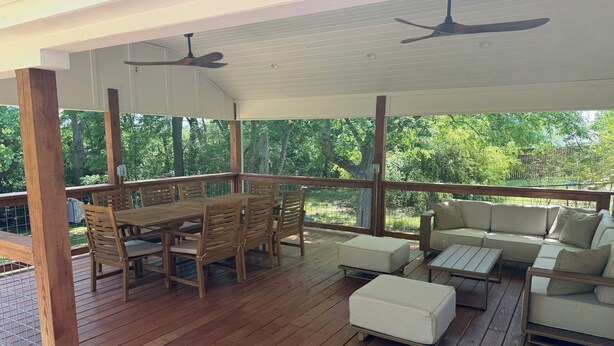 A covered outdoor area with a wooden deck, a dining table with chairs, a sofa, and a ceiling fan.