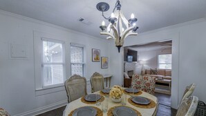 A dining room with a round table set for four, surrounded by patterned chairs. A chandelier hangs from the ceiling, and there are two windows with blinds.