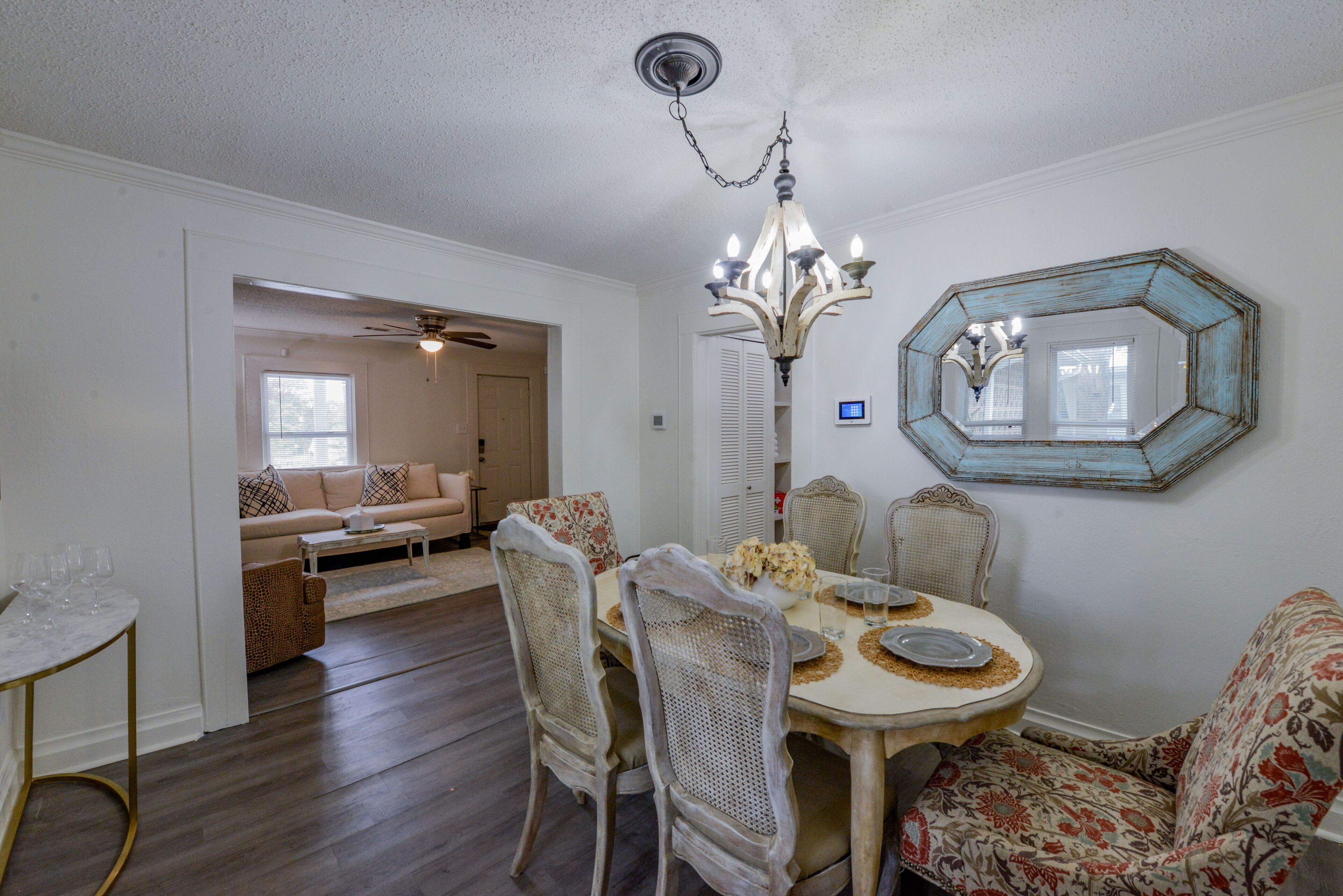 A dining area with a round table, chairs, and a chandelier. There is a sofa and a window in the background.